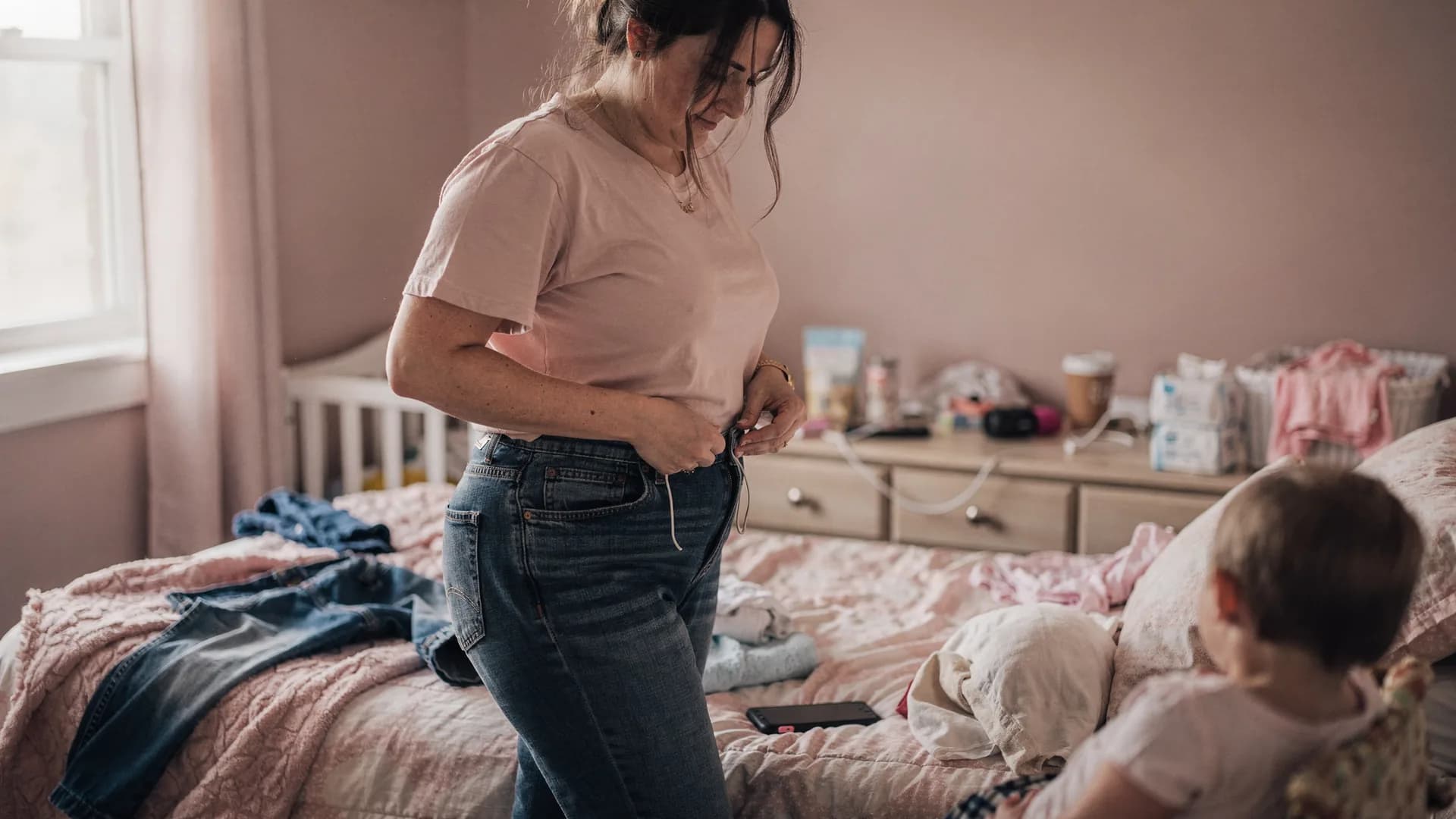 A woman fastens a comfortable pair of jeans in a bedroom with another pair on the bed.