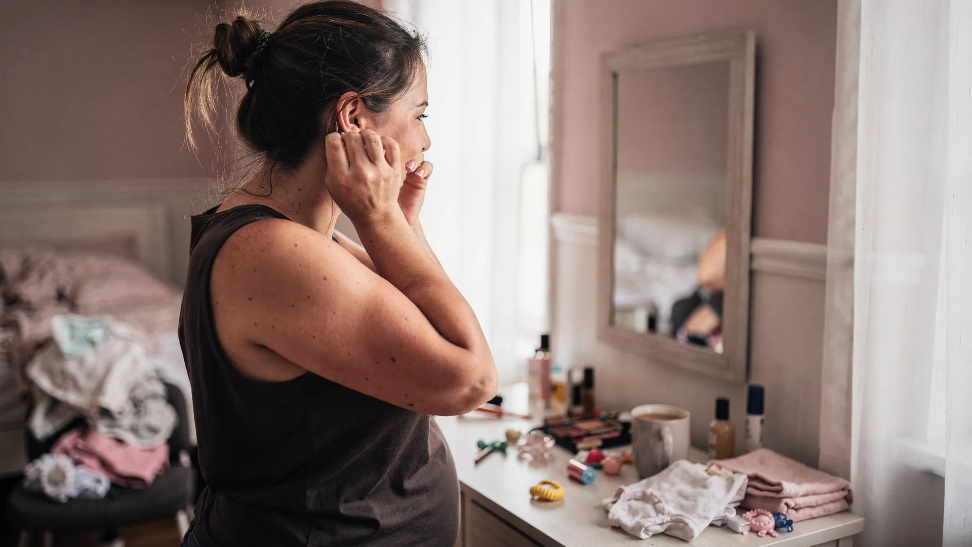 A woman clasps earrings in a bedroom mirror with baby clothes and makeup nearby.