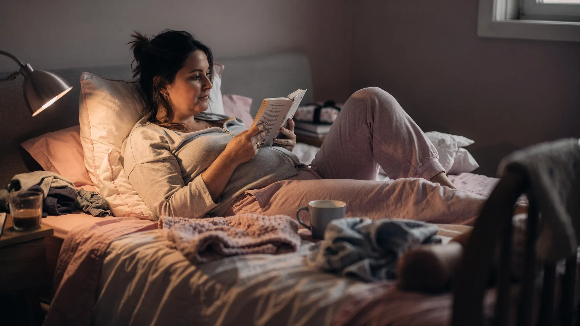 A woman reads alone in a dim bedroom while the other side of the bed is softly out of focus.