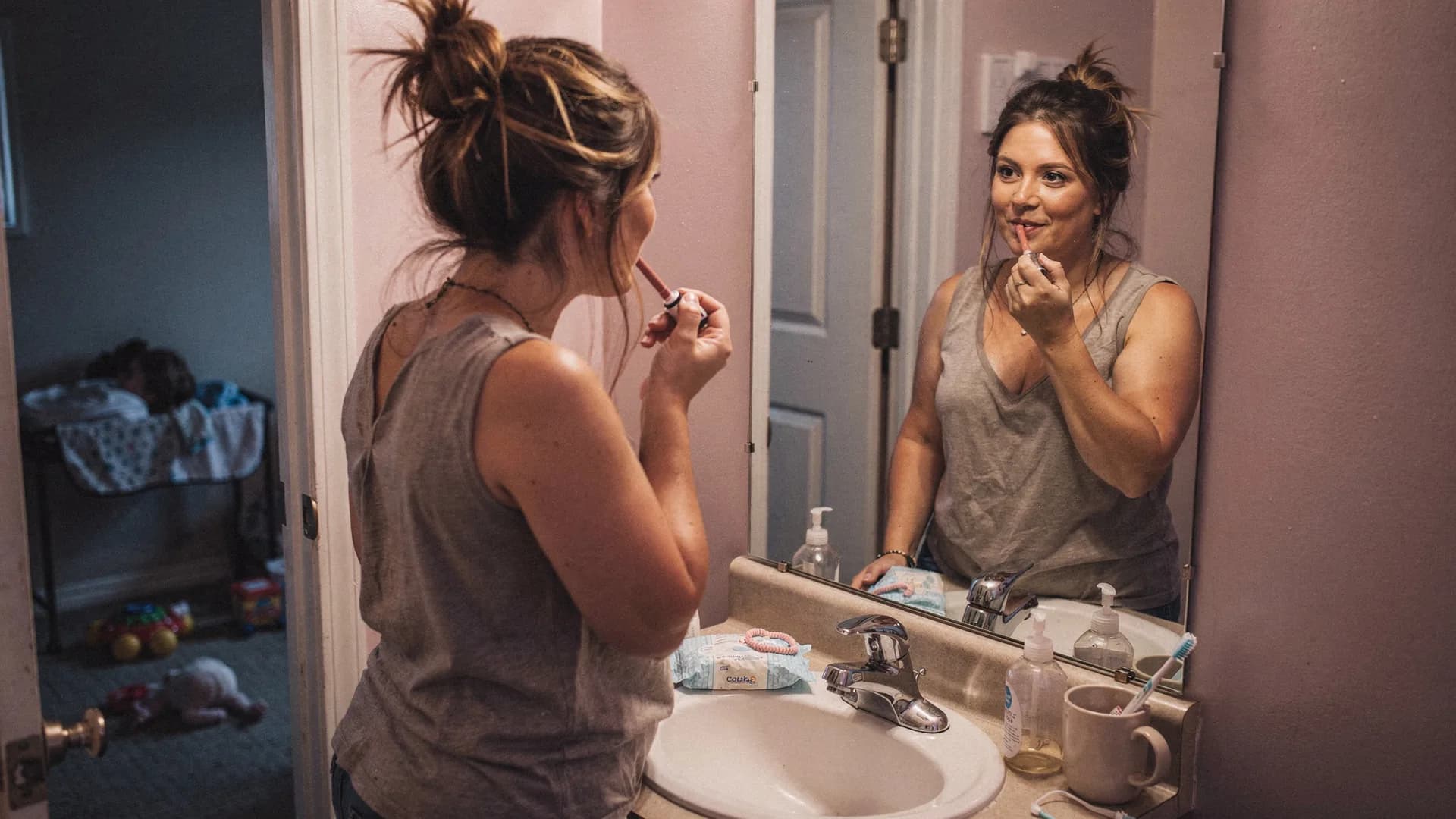 A woman applies lipstick in a small bathroom with baby wipes on the sink.