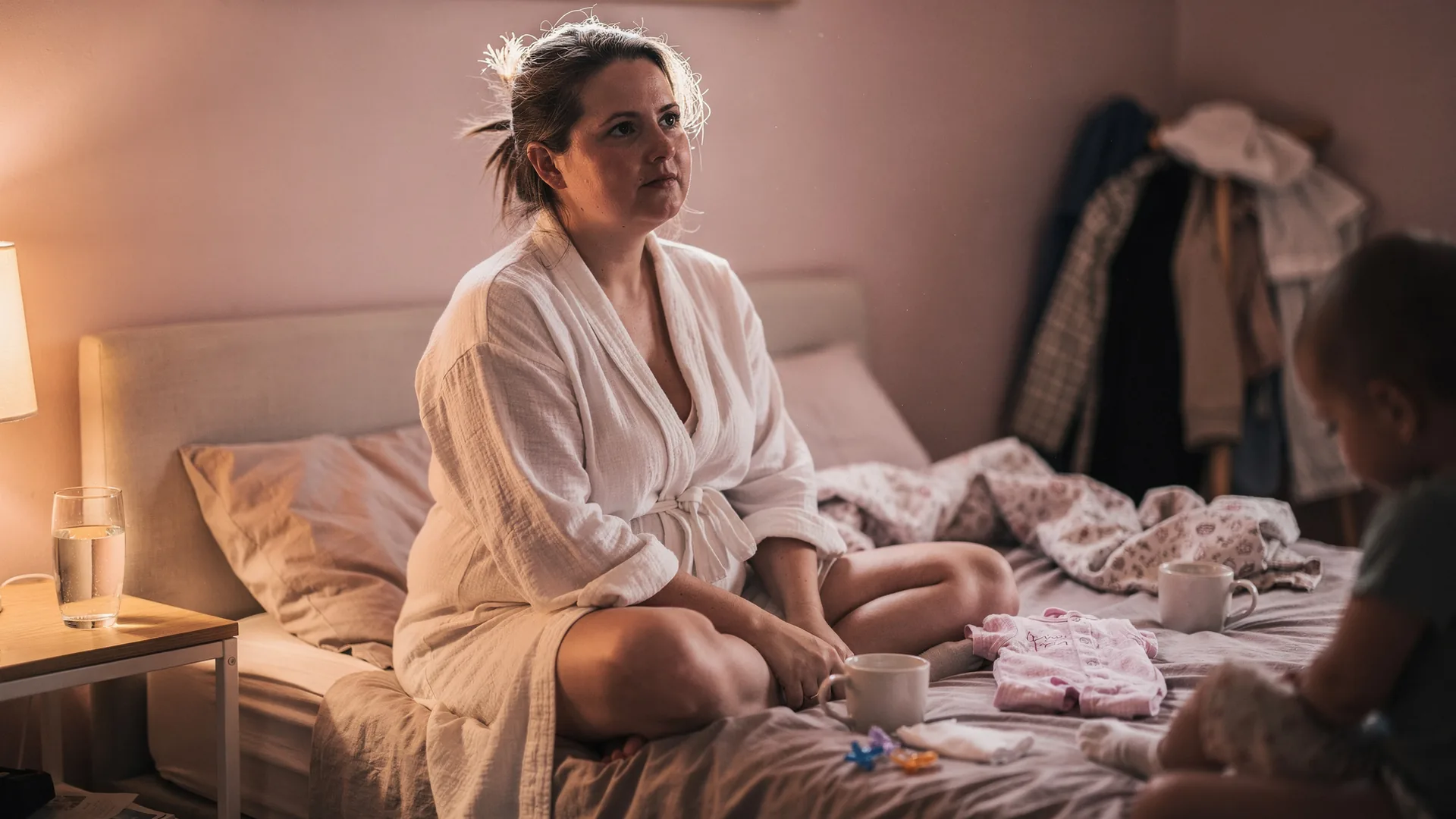 A woman sits thoughtfully on a bed in a robe under soft lamplight.
