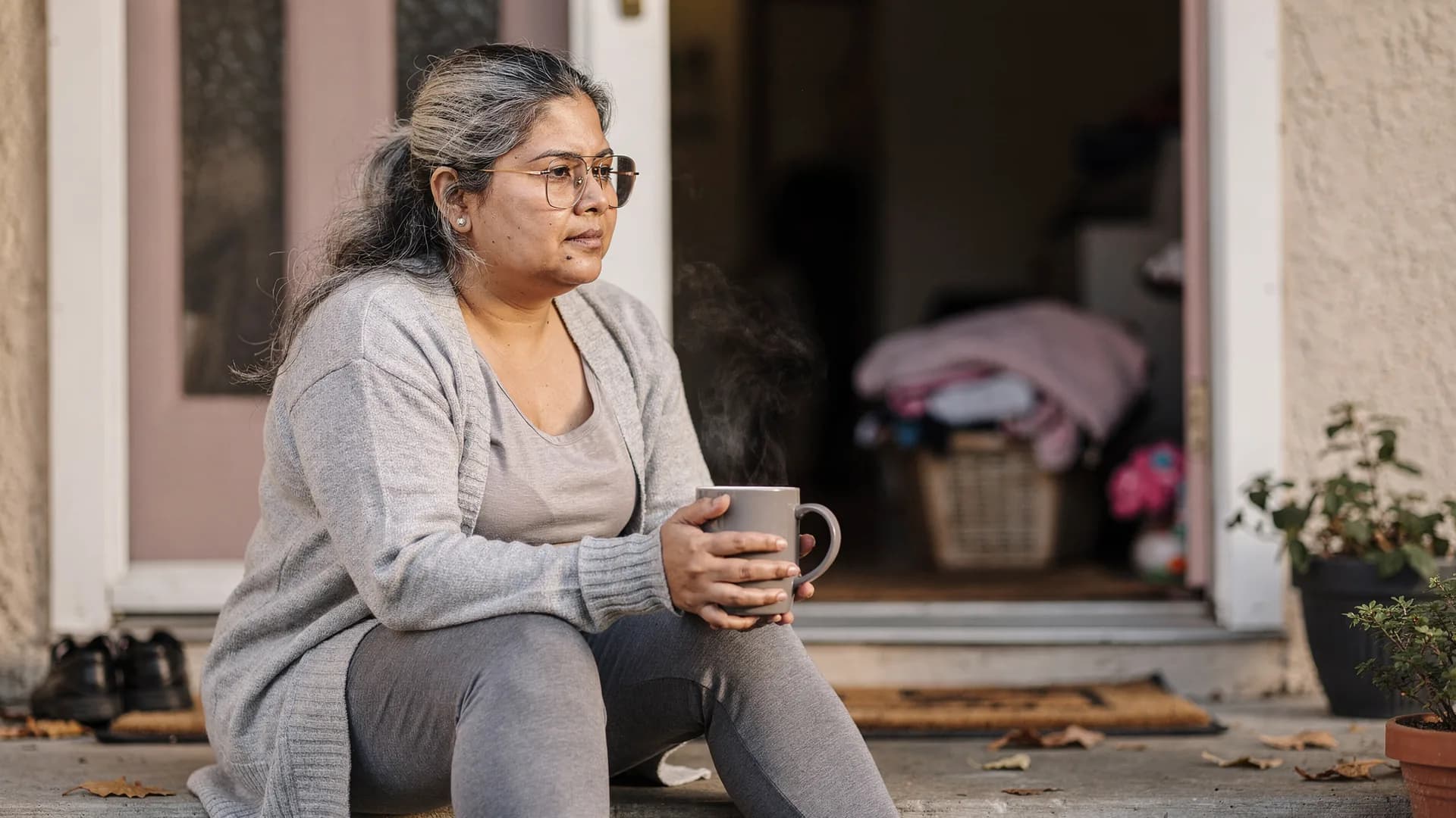 A woman sits alone on a front step with coffee while the door is cracked behind her.