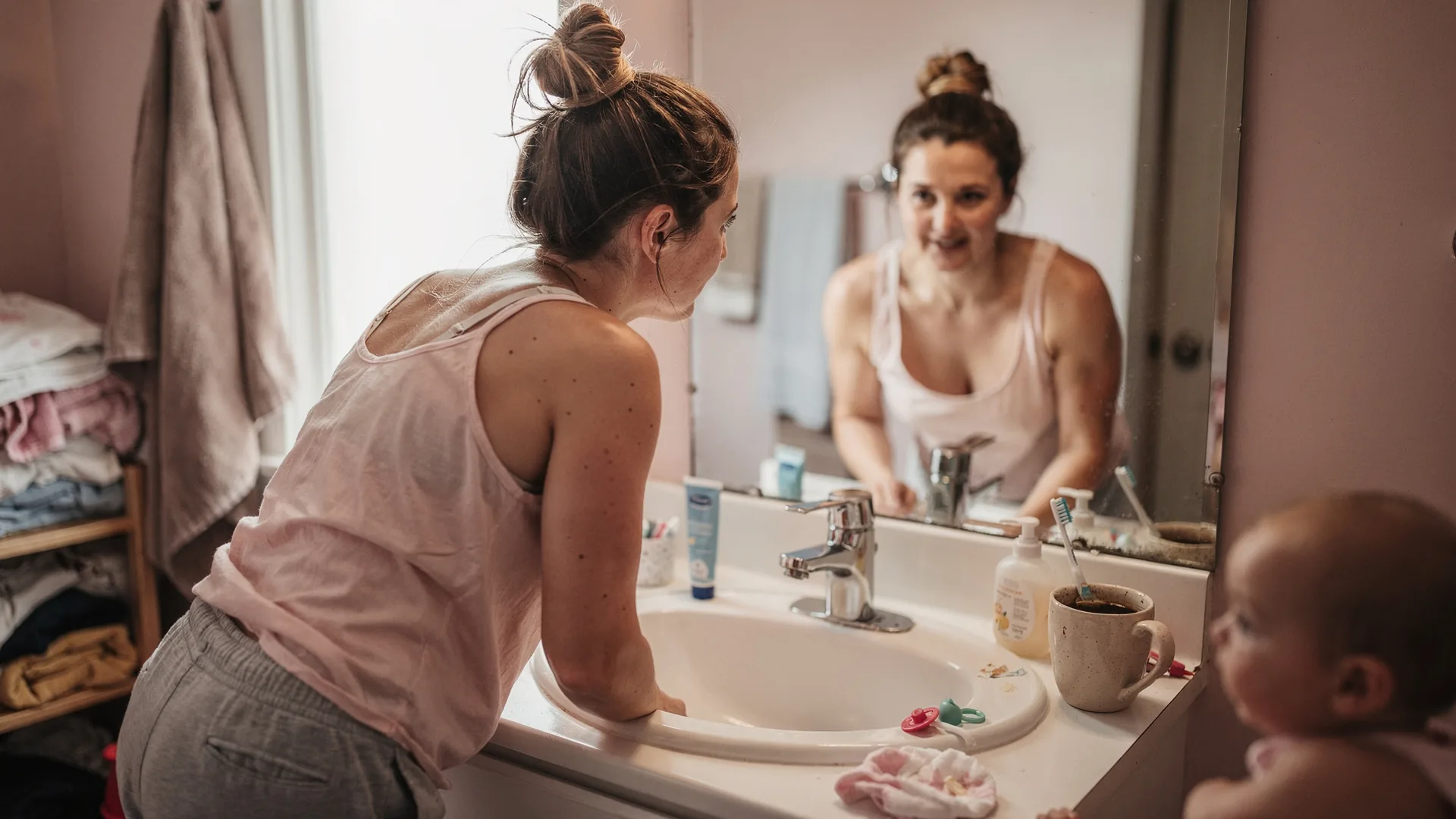 A woman leans over a bathroom sink and looks steadily into the mirror in warm morning light.