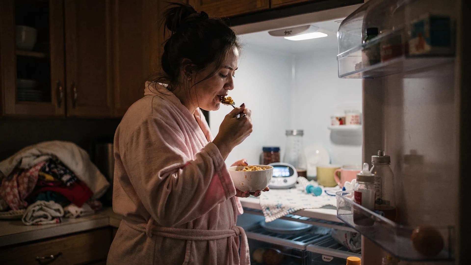A woman in a robe eats cereal at night in a kitchen lit by the open fridge.