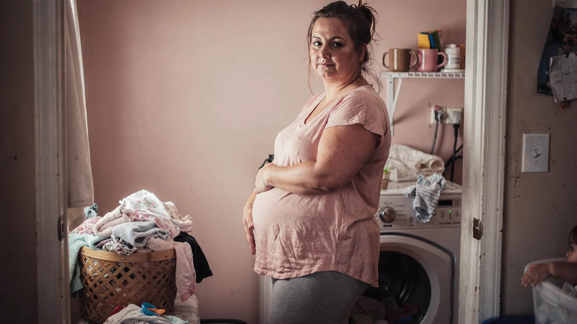 A woman stands calmly in a laundry room doorway beside piles of baby clothes.