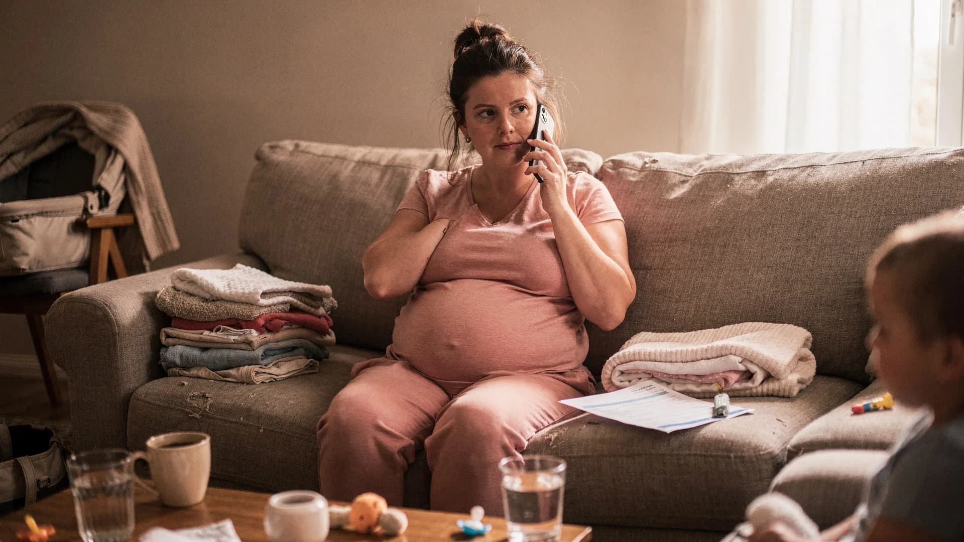 A woman sits on a couch making a phone call with discharge papers beside her.