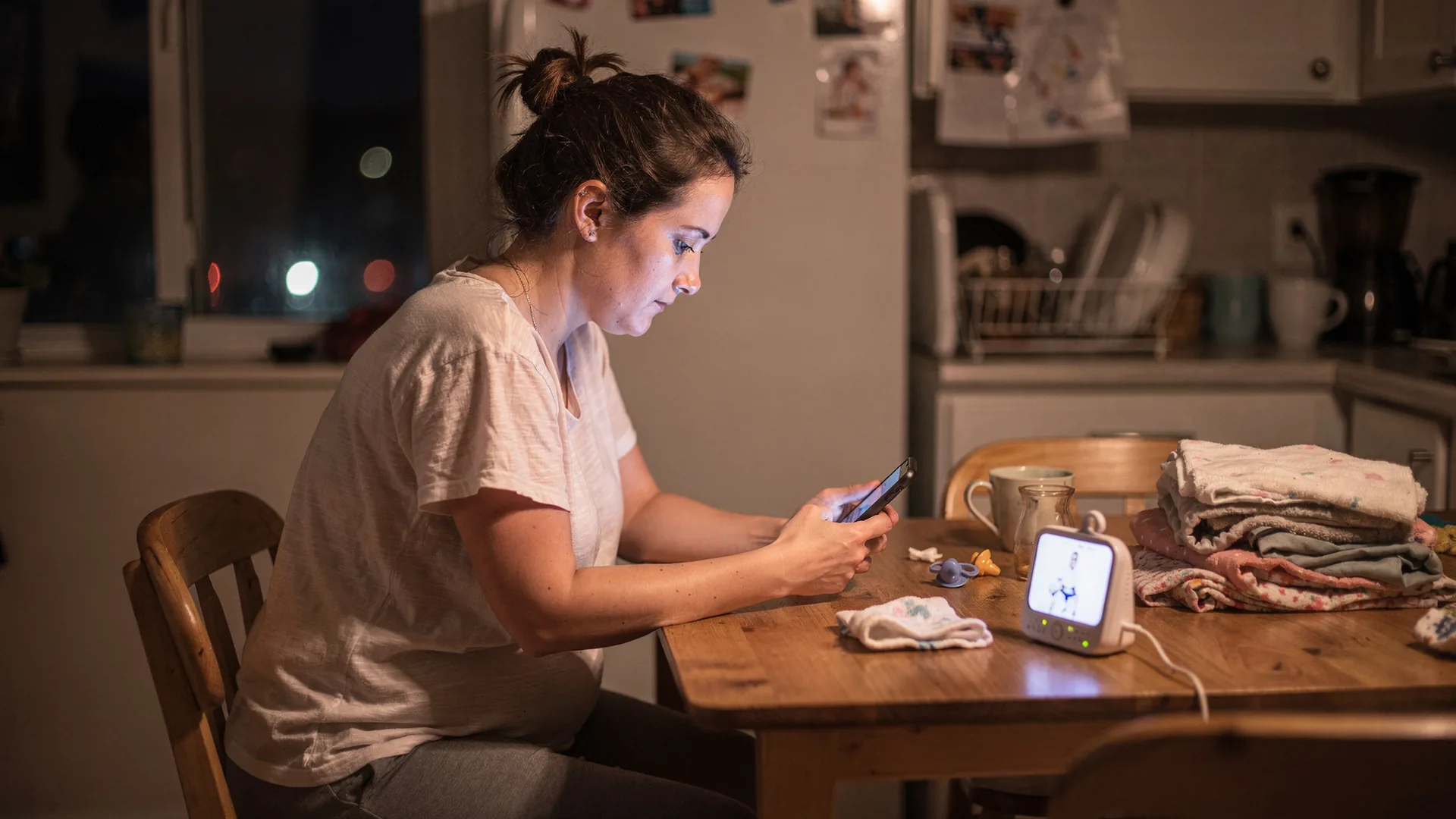 A woman sits alone at a kitchen table at night looking at an old photo on her phone.